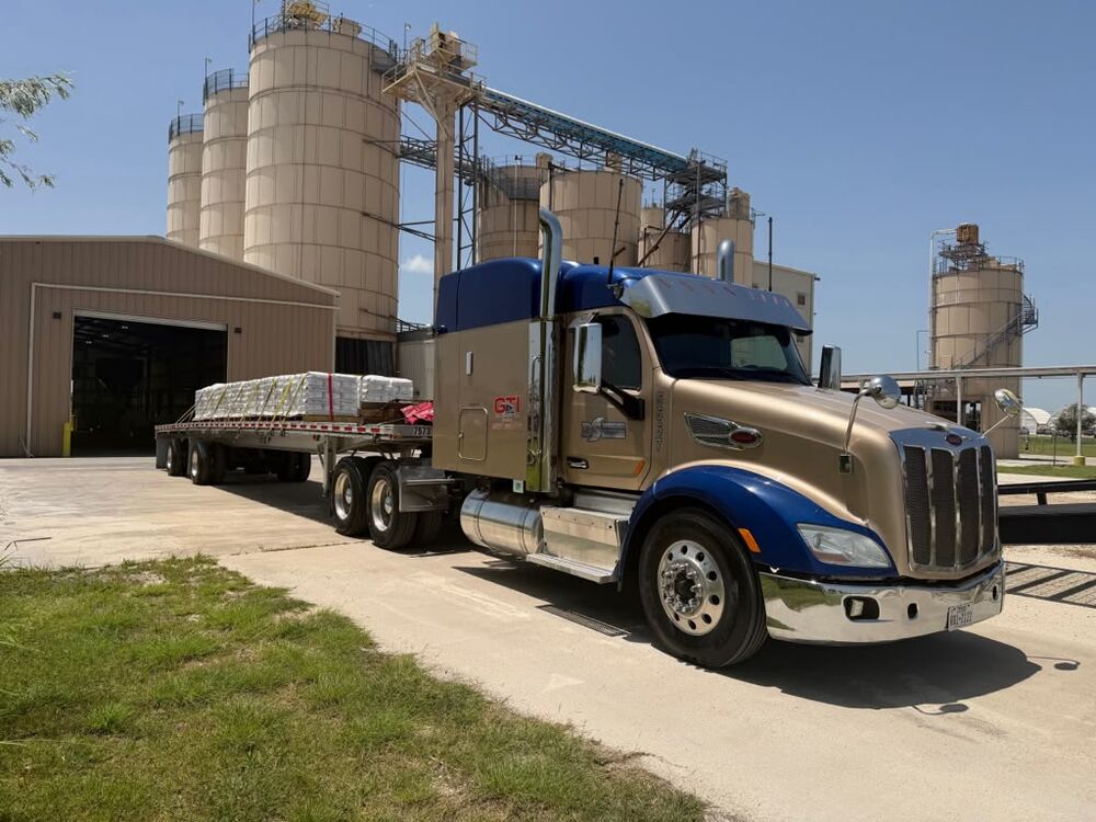 A gold and blue GTI Transport Peterbilt with a flatbed of palletized freight at a cement plant