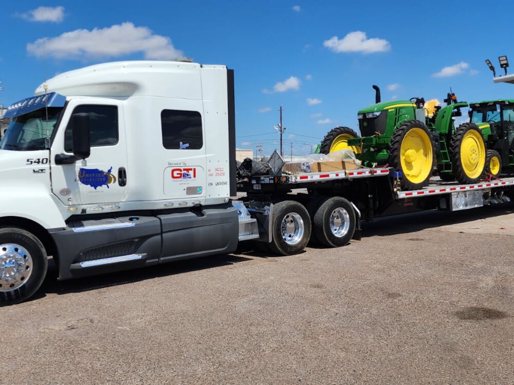 A white GTI Transport tractor hauling John Deere tractors on a step-deck trailer